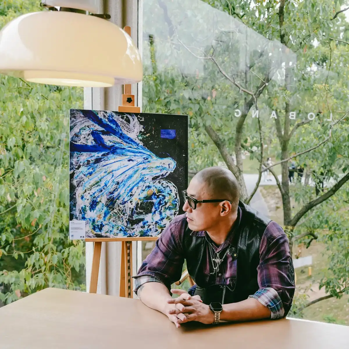 A seated portrait of a man beside a painting and window.