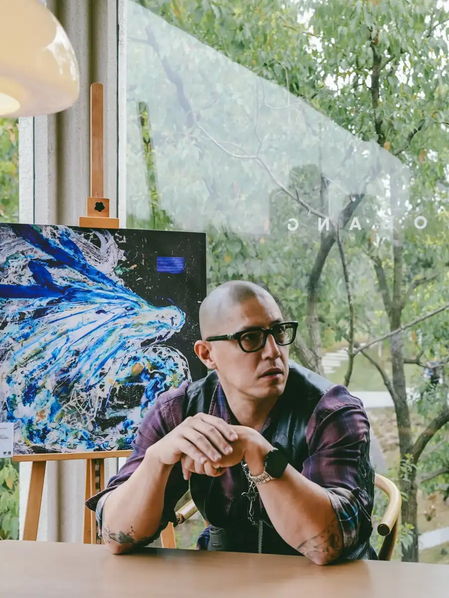 A seated portrait of a man beside a painting and window.