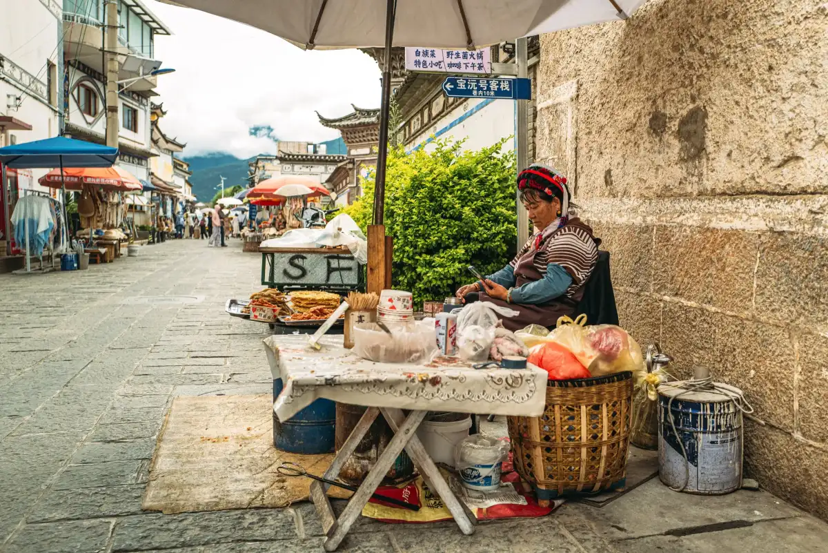 A street vendor seated beside a wall and table.